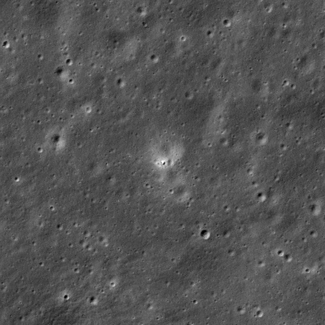 Overhead image of a normal-looking lunar surface lit with a Sun elevation about 40 degrees above the horizon. At the center is a white dot, the Chang'e 6 lander, a few pixels across (about 3 meters), surrounded by a ten-to-thirty meter radius region of lighter-than-normal terrain. The lander is flanked by two craters similar in size to it, and is on the edge of a much more subtle crater about 50 meters wide.